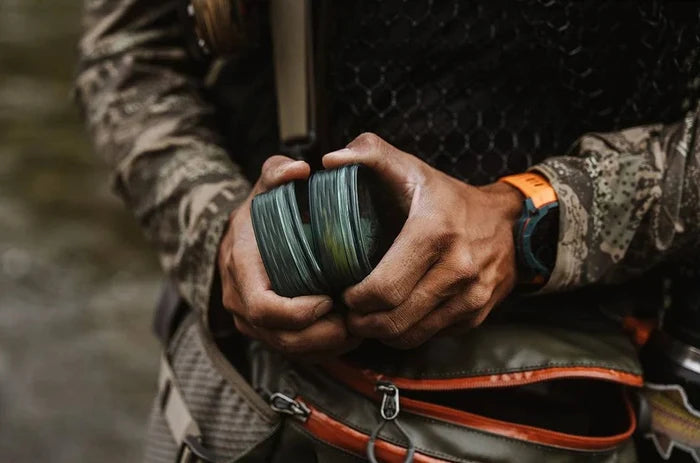 Angler holding a Fishpond Predator Fly Puck with Steelhead flies, wearing camouflage clothing and fishing gear