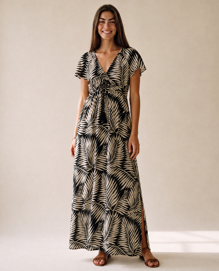 A woman modeling a black and beige palm leaf print maxi dress with short flutter sleeves, a V-neckline, and a tie detail at the waist, standing against a neutral beige studio backdrop.