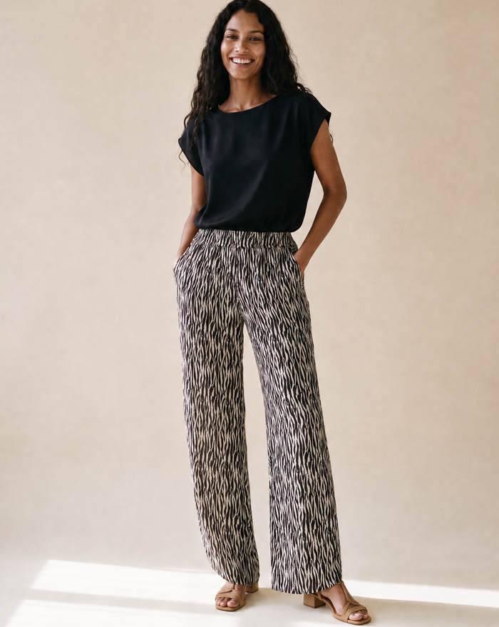 A woman stands in a studio wearing a black short-sleeve top and high-waisted, wide-leg trousers with an abstract black, white, and gray pattern. She is wearing tan heeled sandals against a warm beige background.