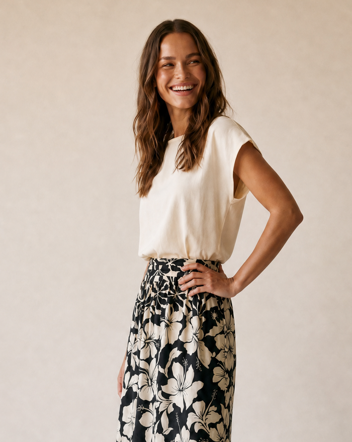 A woman modeling a cream short-sleeve top and a black maxi skirt with a large cream floral print, standing against a warm beige studio backdrop.