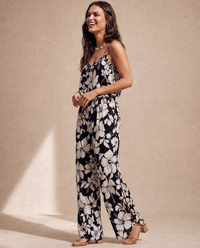A woman modeling a sleeveless black jumpsuit with a large white floral print, standing against a textured beige studio backdrop.