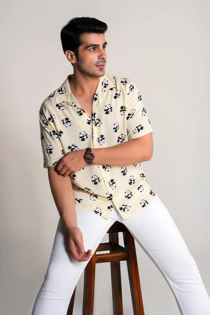 Man wearing panda print yellow oversized shirt and white pants sitting on wooden stool